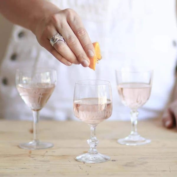 A hand garnishes a glass of rosé wine with an orange peel, with two other glasses nearby.