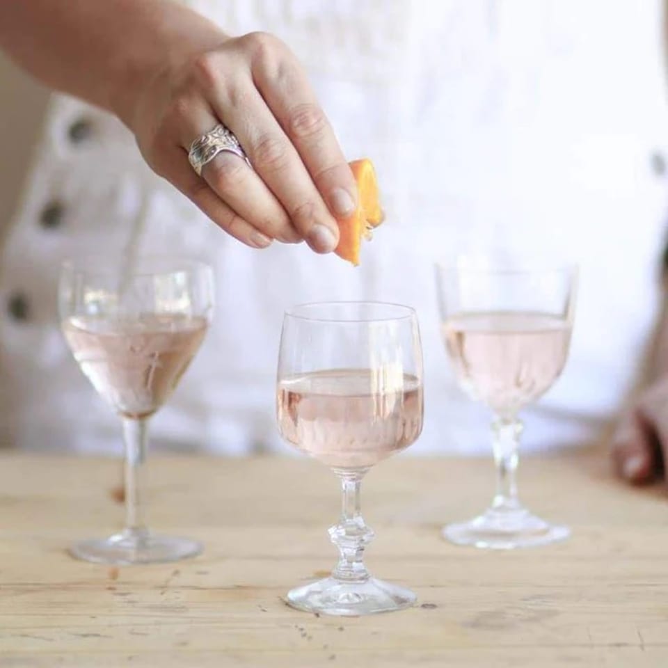 A hand garnishes a glass of rosé wine with an orange peel, with two other glasses nearby.