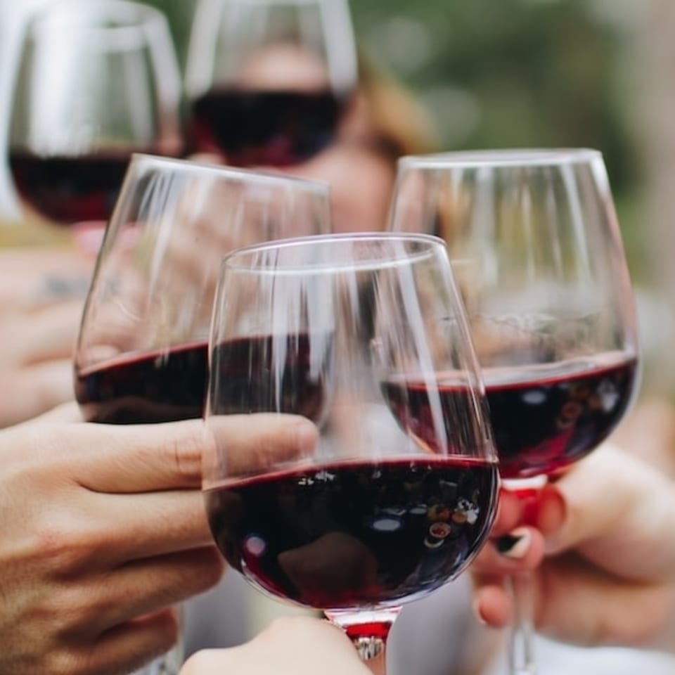 Multiple hands holding up wine glasses filled with red wine, toasting together.