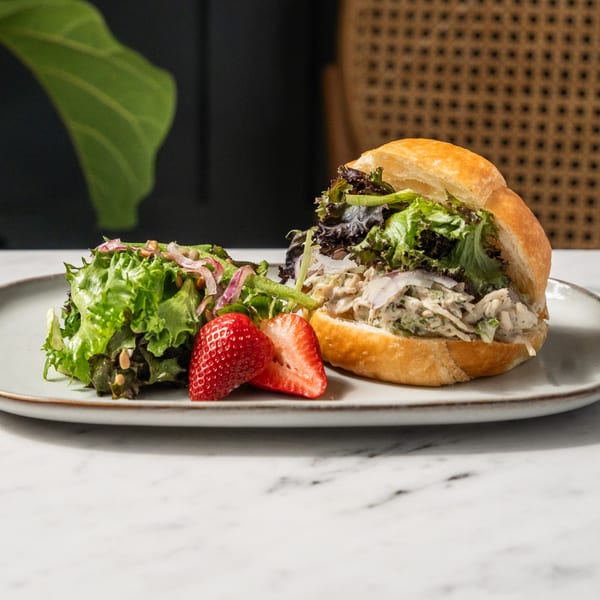 A chicken salad sandwich on a croissant with a side salad and strawberries on a white plate.