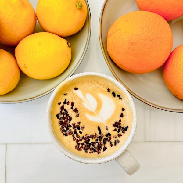 Overhead view of a latte with cocoa nibs and latte art, flanked by plates of lemons and oranges.