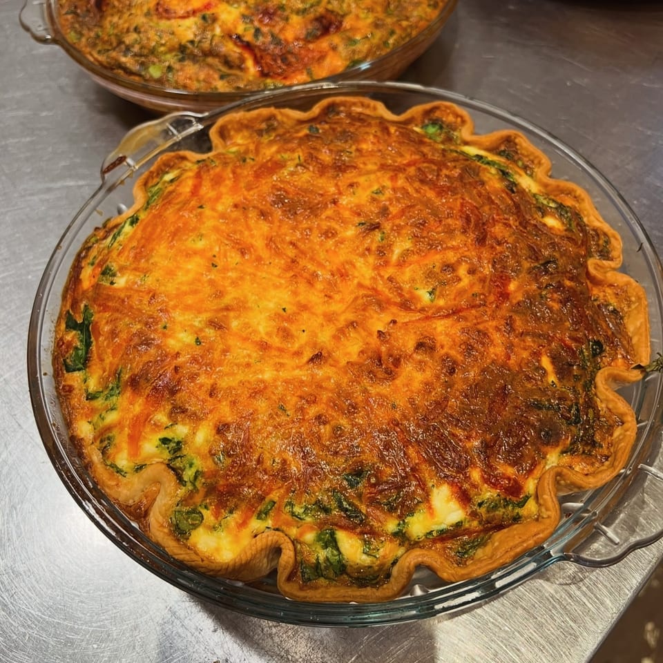 Two golden-brown quiches with flaky crusts in glass pie dishes on a metal surface.