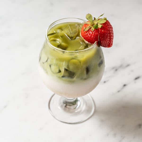 Iced matcha latte with a strawberry garnish in a stemmed glass on a marble surface.