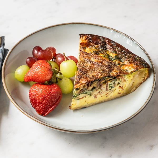 A slice of quiche with a side of red and green grapes and two strawberries on a plate.