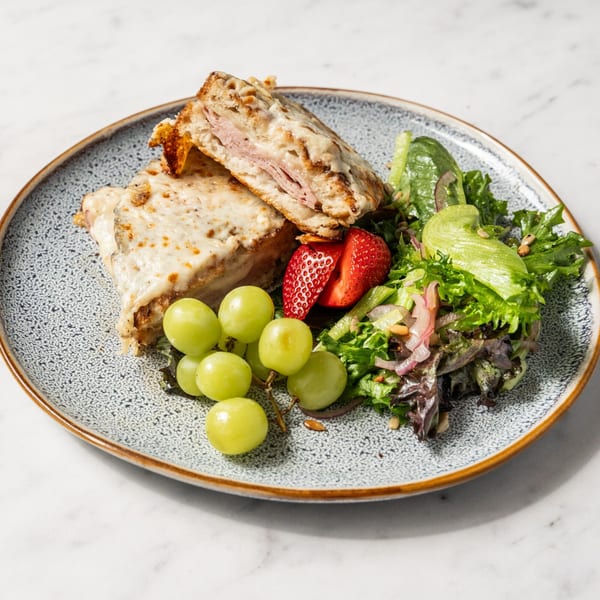 Croque Monsieur sandwich with a side salad, green grapes, and a sliced strawberry on a speckled plate.